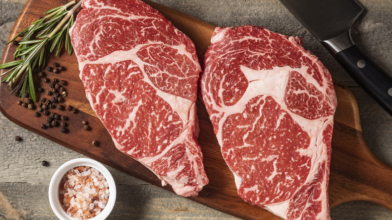 Two raw marbled steaks on a wooden cutting board surrounded by seasonings