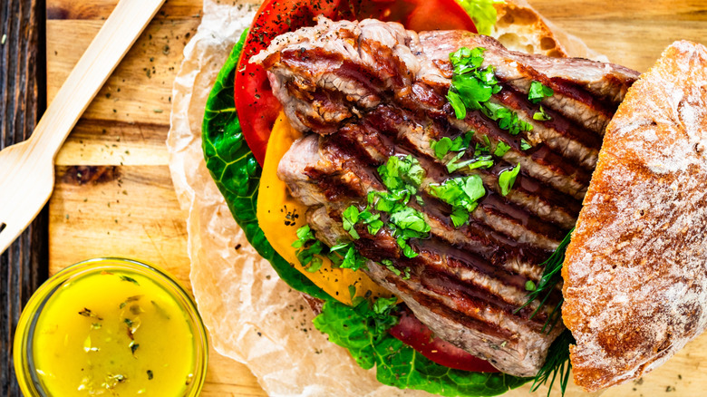 A steak sandwich on a wooden board with a side of herby dressing