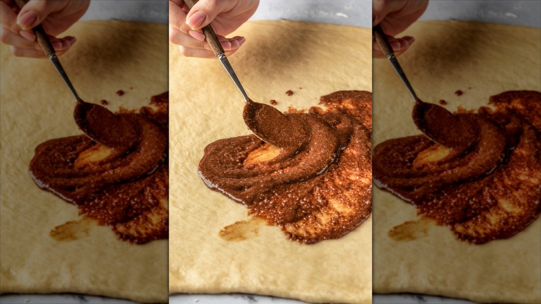A hand spooning cinnamon filling onto a large piece of rolled dough.