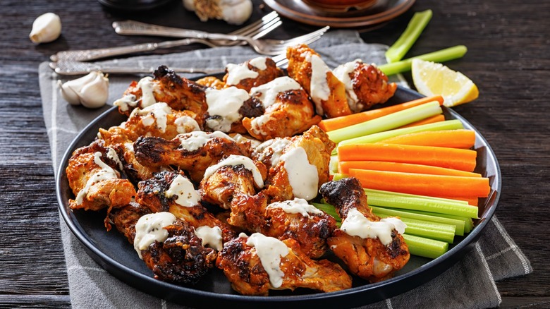 Grilled chicken wings with Alabama white sauce on a plate with celery, and carrots sticks on a dark wooden table