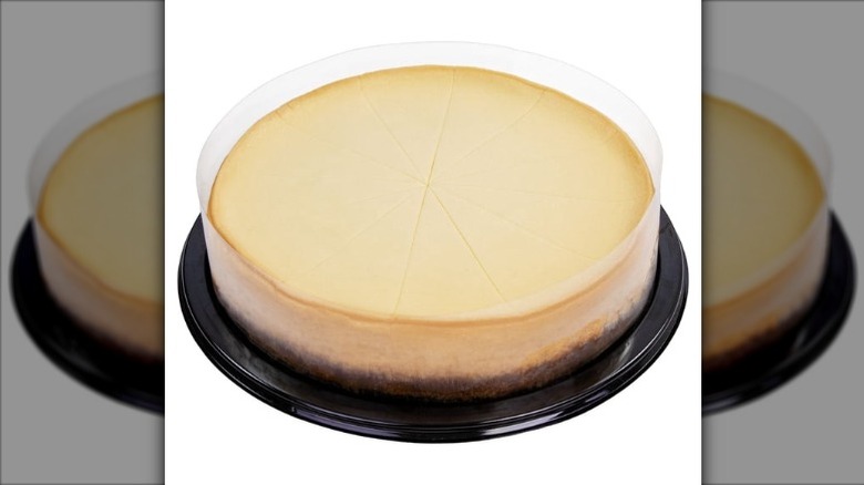 A plain cheesecake on a black plastic base covered in a clear plastic dome