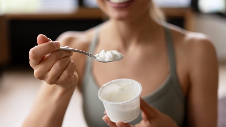 Close-up of a smiling sportswoman holding a spoon with fresh yogurt and a yogurt pot in her hands after a workout, enjoying a healthy protein-rich snack. Food concept