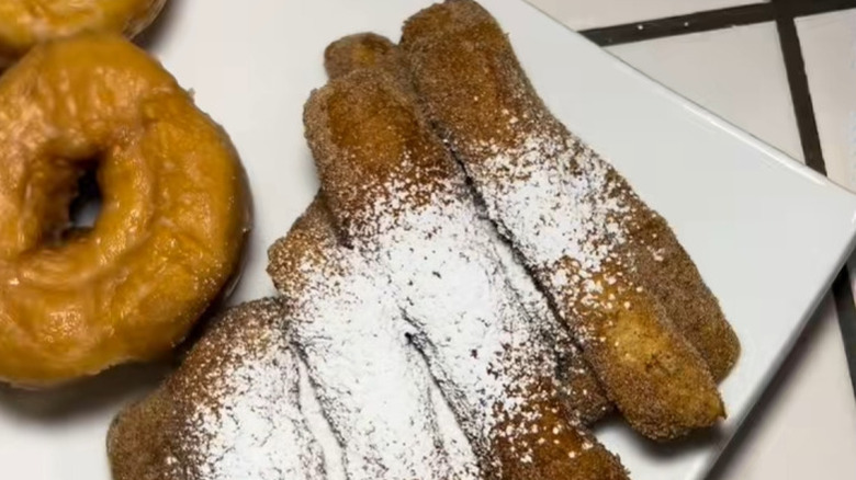 Homemade churros created from pizza dough and sprinkled with icing sugar rest on a plate beside donuts.
