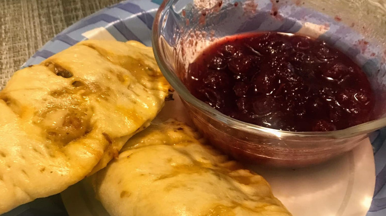 Two empanadas sit on a paper plate, a bowl of red dipping sauce to the right of them.