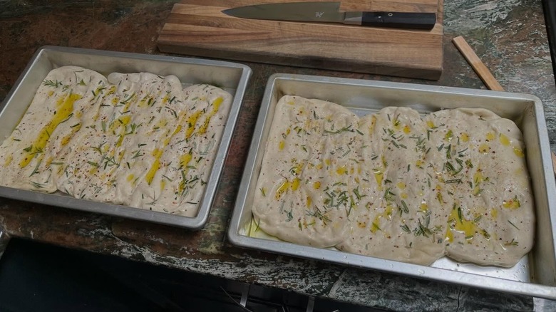 Two metal trays of uncooked Focaccia rest side by side on a wooden table.