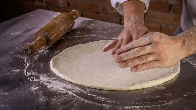 A chef kneads pizza dough on a metal counter. A rolling pin sits to the left side.