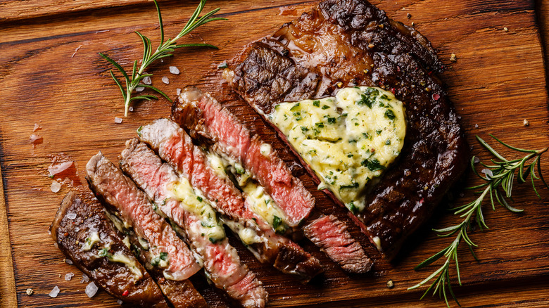 A cooked piece of steak that is sliced and smothered in butter with rosemary sprigs around it.