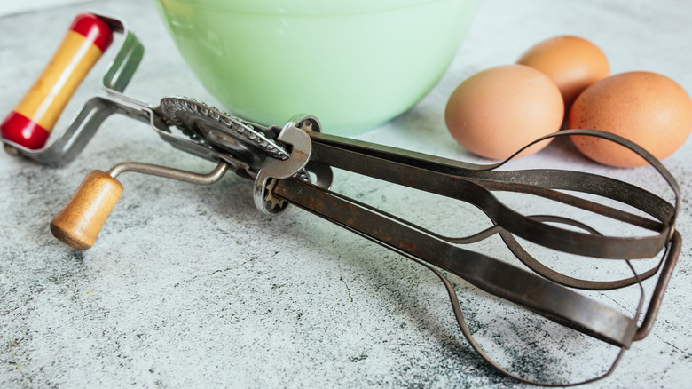 A hand-held egg beater rests on a table. A green bowl and three brown eggs sit in the background
