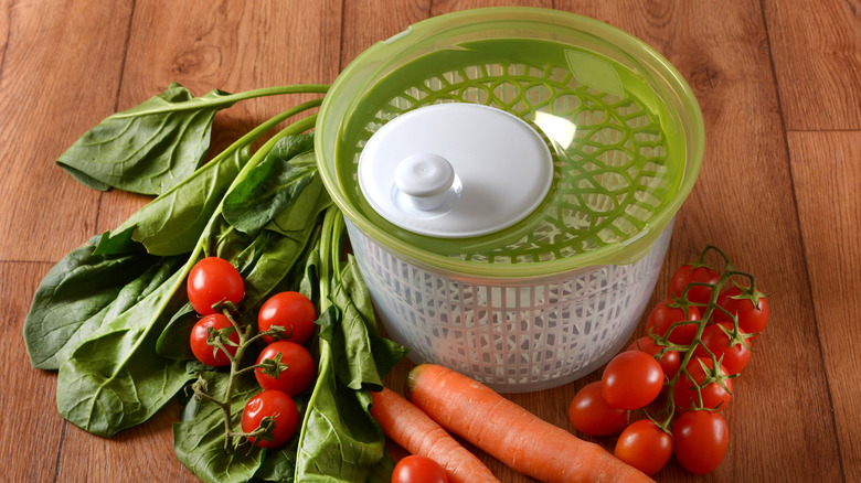 A salad spinner sits on a wooden floor, vegetables surrounding it