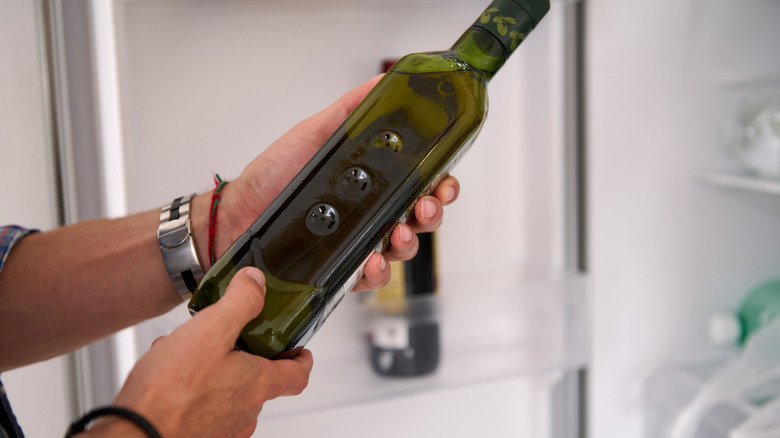 Person's hands holding a dark glass bottle of oil in front of a fridge