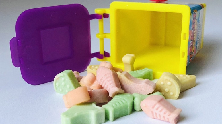 A can of Garbage Can-dy tipped over to spill colorful candies shaped like common trash items onto a white surface