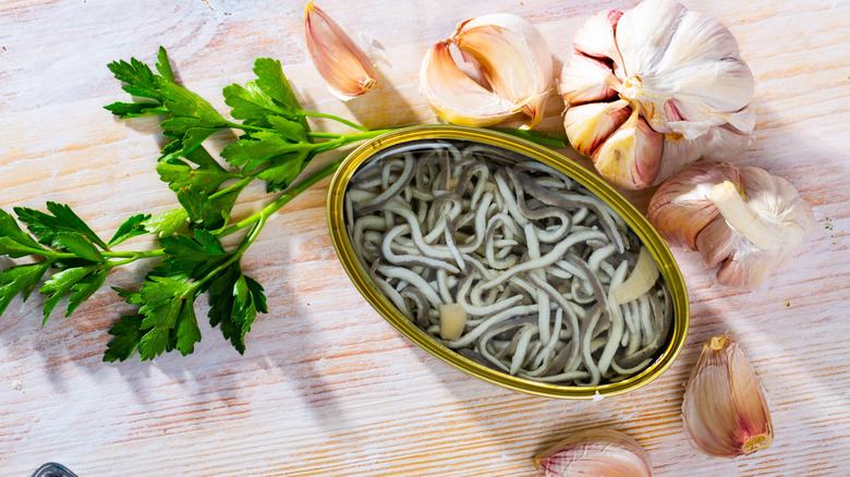 Top view of open tin of canned baby eels surrounded by fresh parsley and unpeeled garlic cloves