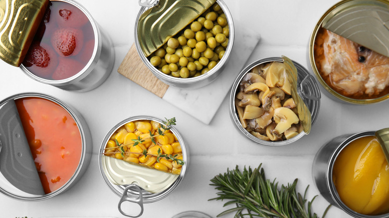 Top down view of opened assorted canned good, like mushrooms, peaches, peas, and corn