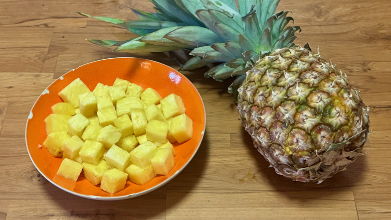 Cubes of pineapple in an orange bowl next to a whole pineapple on a wooden table