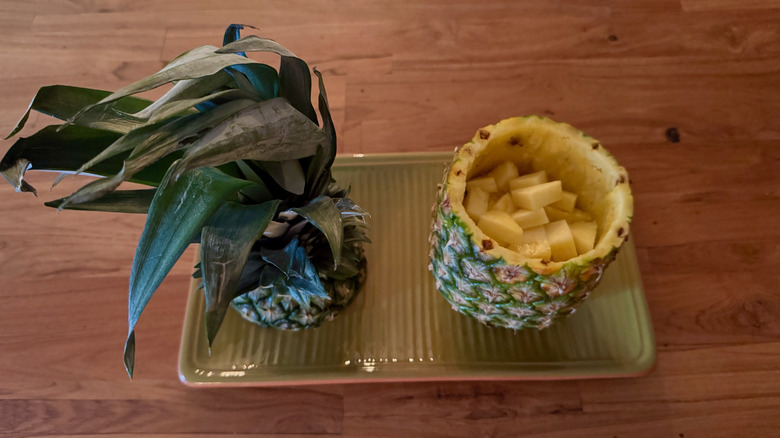 Small wedges of pineapple in a pineapple bowl on a green platter with the crown of the pineapple next to it