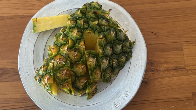 A stick of pineapple on a white ceramic plate, pulled from the back of a half pineapple with a checkered slicing pattern
