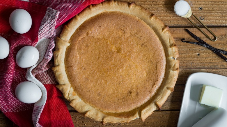 Chess pie on wooden table alongside a teaspoon of sugar, vanilla, a pat of butter, and several whole eggs