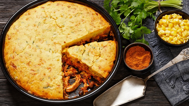 Chicken Tamale Pie consisting of seasoned shredded chicken, salsa sauce, black beans, cheese, and a cornbread crust in a baking dish on dark wood table