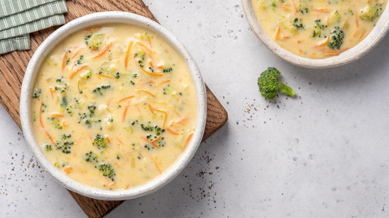 Two bowls of broccoli cheddar soup sitting on a counter with a stray piece of broccoli.