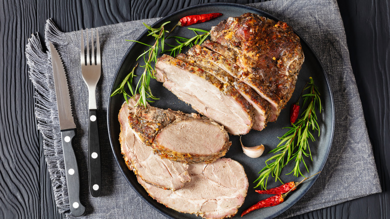 Roast Pork Neck cut on a dark grey plate on black wooden table with dried red pepper, rosemary, garlic, fork and knife, horizontal view from above
