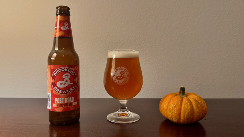 A glass bottle of Brooklyn Brewery Post Road beer next to a full beer glass and a small pumpkin.