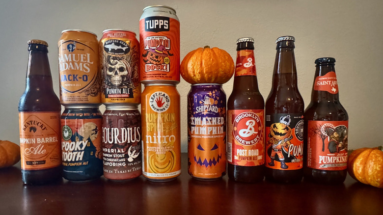 11 bottles and cans of pumpkin beer lined up on a wooden table with small pumpkins, including brands like Samuel Adams, Shipyard, and Tupps