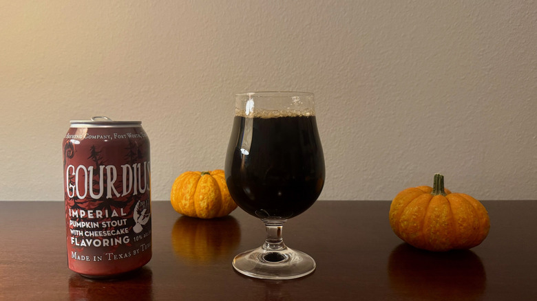 A goblet of dark beer next to a brown can of Martin House Brewing Gourdius and two small pumpkins