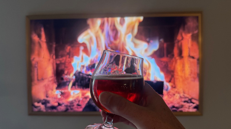 A glass of pumpkin beer held in front of a fireplace playing on a TV screen.