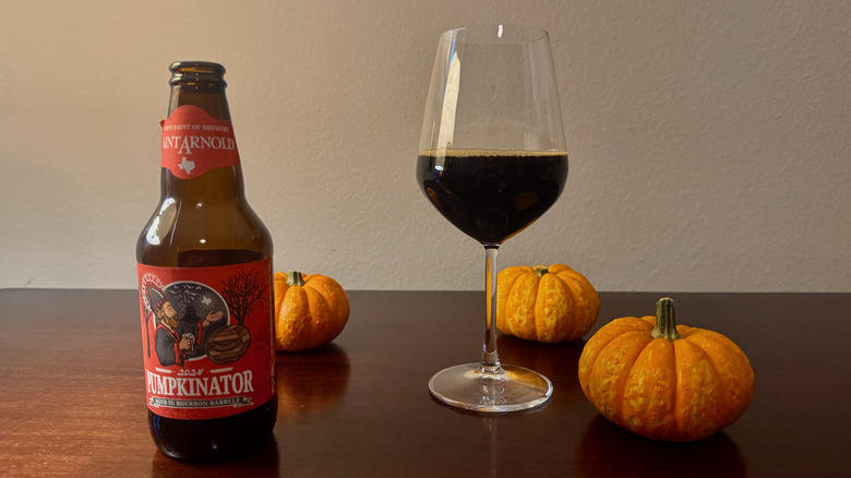 A bottle of Saint Arnold Pumpkinator next to a wine glass half-filled with dark beer and three small pumpkins.
