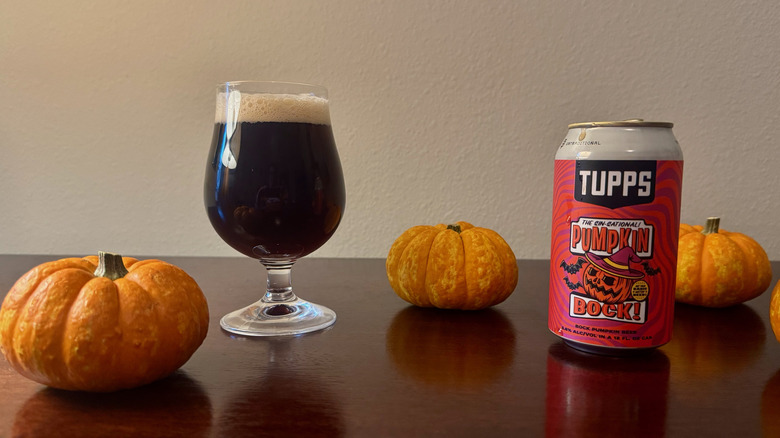 A goblet of dark beer next to can of Tupps Pumpkin Bock and three small pumpkins.
