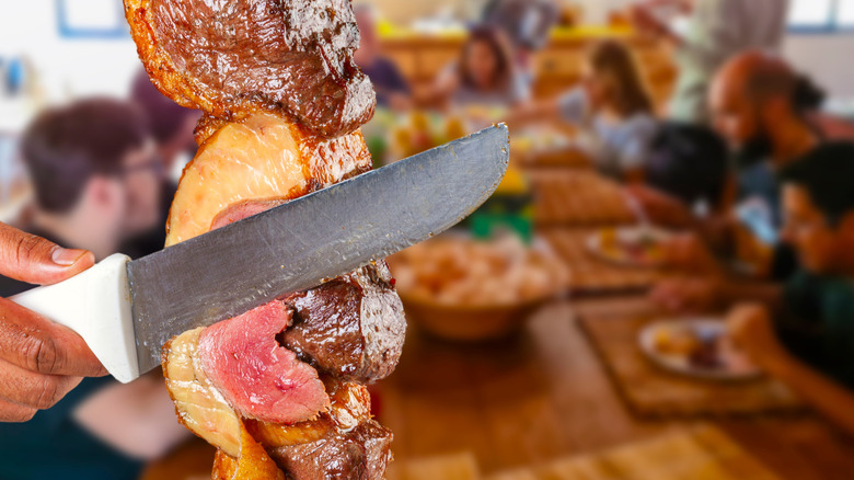 A close up of meat being cut from a skewer as a table waits in the background.