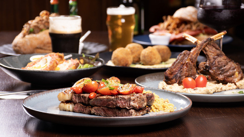 Several different plates of meat and sides, are arranged on a wooden table.