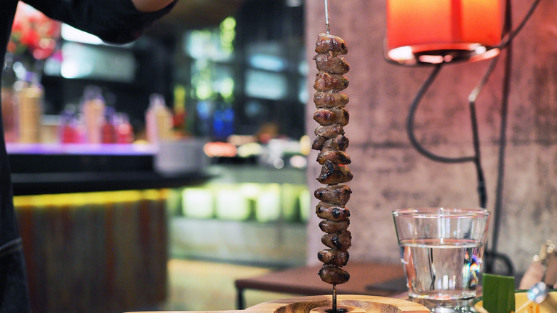 A tall skewer of meat rests upright on a wooden resturant table, a glass of water to the right.