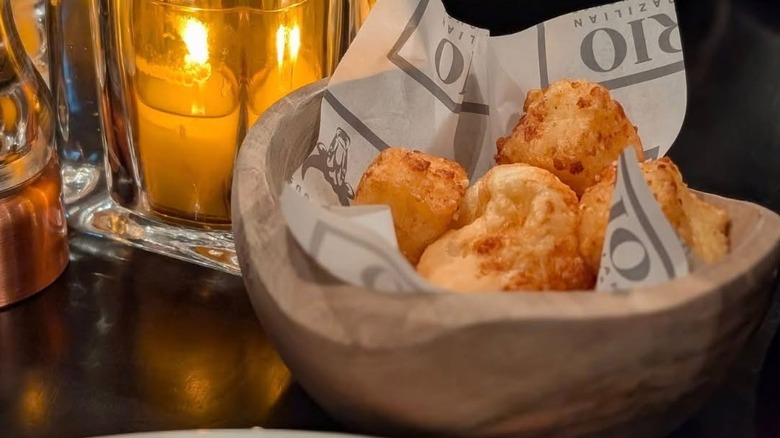 A bowl full of pão de queijo rests on a resturant table, a lit candle in the background.