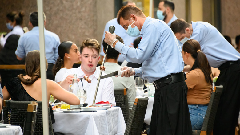 A couple sit at a table while a gaucho serves meat to them from a skewer.