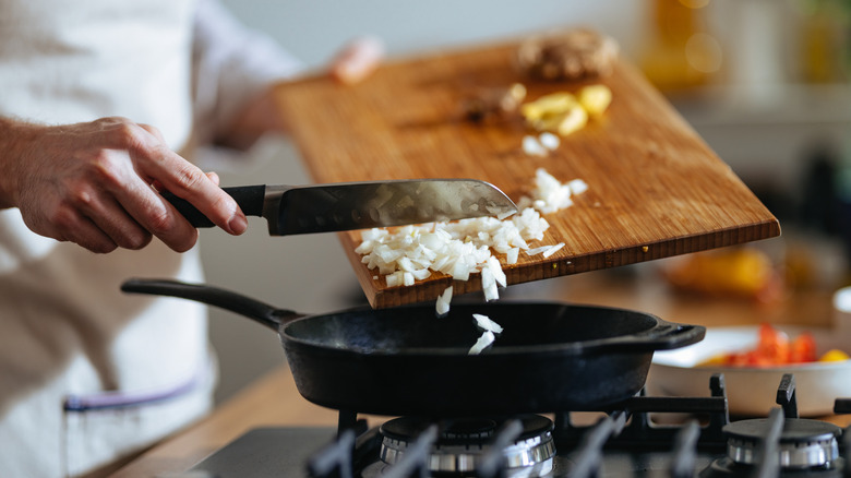 Person scraping chopped onions off cutting board with knife into black frying pan