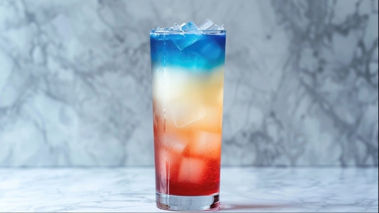 A red, white, and blue layered cocktail in a tall glass with ice cubes sitting on a kitchen counter