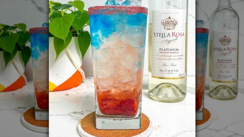 A red, white, and blue layered cocktail in a tall glass of ice shown sitting between a potted plant and a bottle of Stella Rosa vanilla-flavored white wine