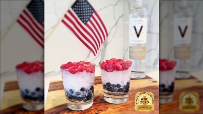 Two drinks made with layers of berries shown with an American flag and a bottle of vodka in the background.