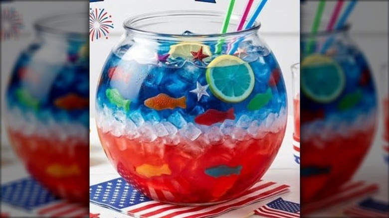 A red, white, and blue layered cocktail in a fishbowl filled with crushed ice and garnished with gummy fish and lemon slices.