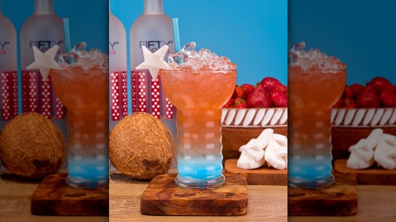 A red, white, and blue layered cocktail in a tall glass of ice pictured with a coconut, chunks of coconut, strawberries, and two vodka bottles