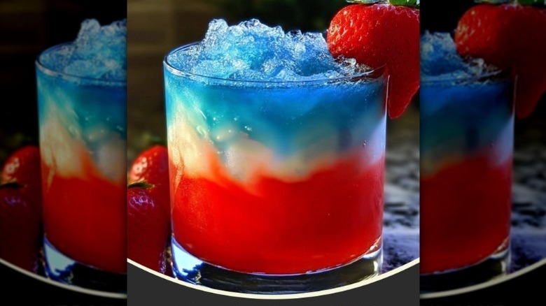 A red, white, and blue layered cocktail in a rocks glass filled with crushed ice and garnished with a strawberry