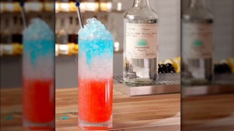 A red, white, and blue layered cocktail in a tall glass of ice sitting on a kitchen counter in front of a bottle of tequila