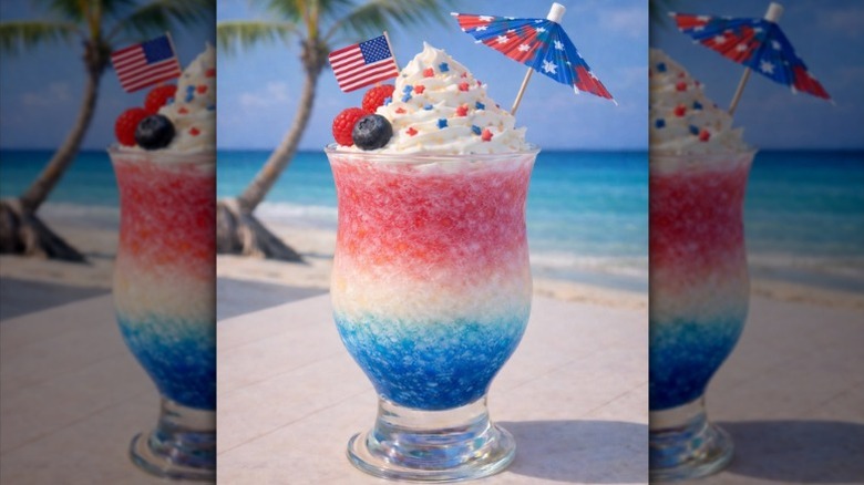 A red, white, and blue layered frozen drink in a hurricane glass garnished with a paper flag, a paper umbrella, berries, whipped cream, and sprinkles