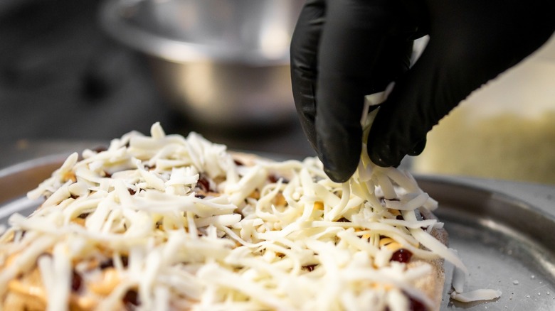 A gloved hand sprinkles shredded cheese onto a pizza