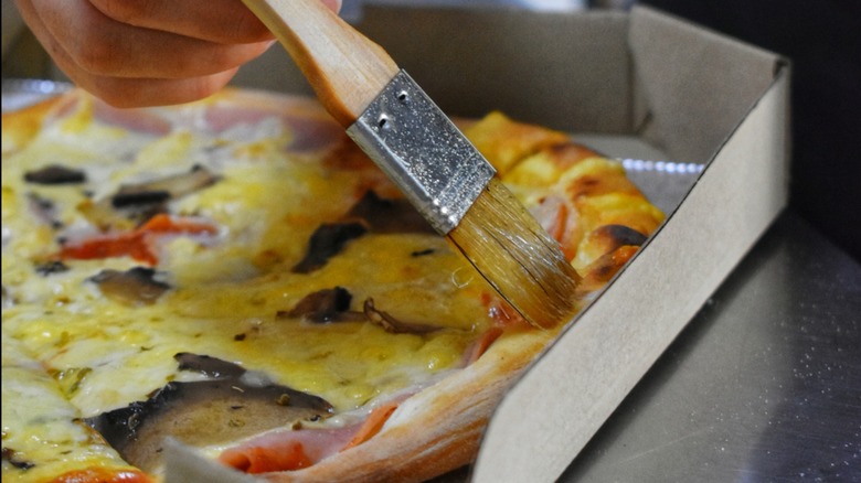 A hand shown adding olive oil to a pizza crust with a brush