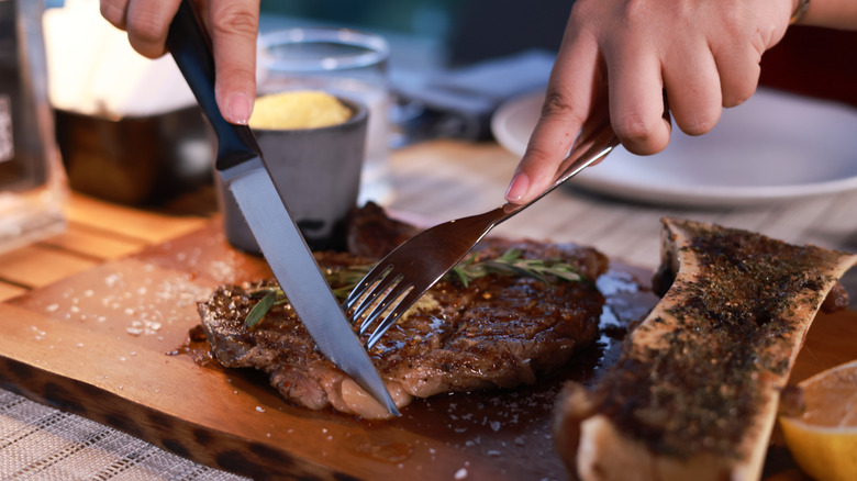 Two hands use knife and fork to cut into a steak on wooden board.