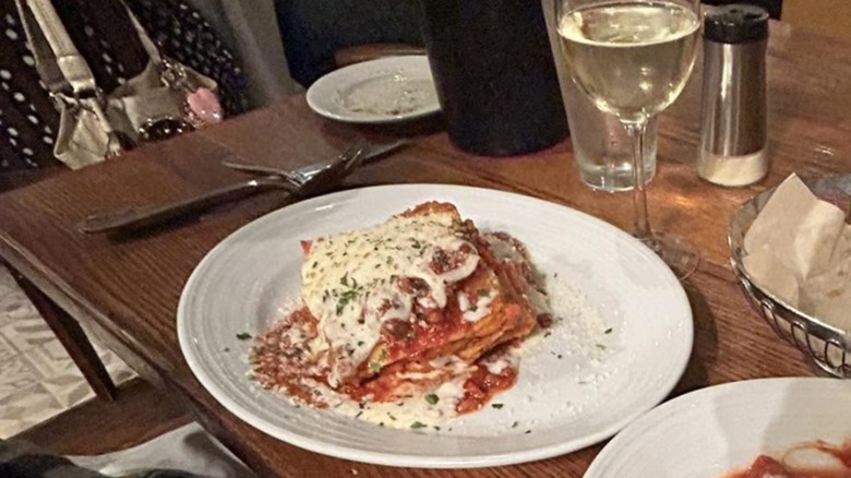 Slice of Biaggi's Ristorante Italiano Lasagna Bolognese on a white plate on wooden table with glass of white wine