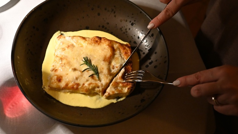 Hands shown eating lasagna with béchamel sauce in a black plate in a restaurant
