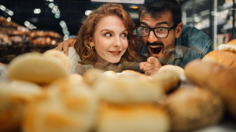 Young couple pointing and choosing fresh bread from bakery display in supermarket, smelling the bread
